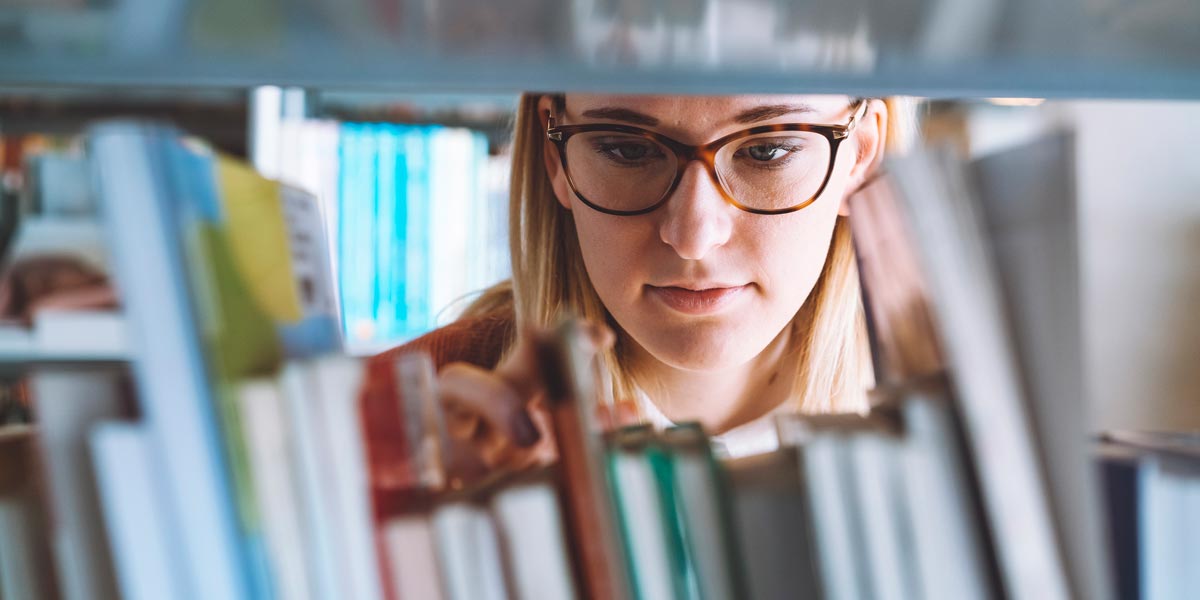 A woman wearing glasses searches for a book on a shelf.
