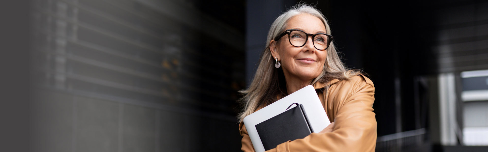 An older woman with gray hair is wearing black-rimmed glasses and a brown leather jacket. She looks into the distance, smiling, while clutching a laptop and notebook.