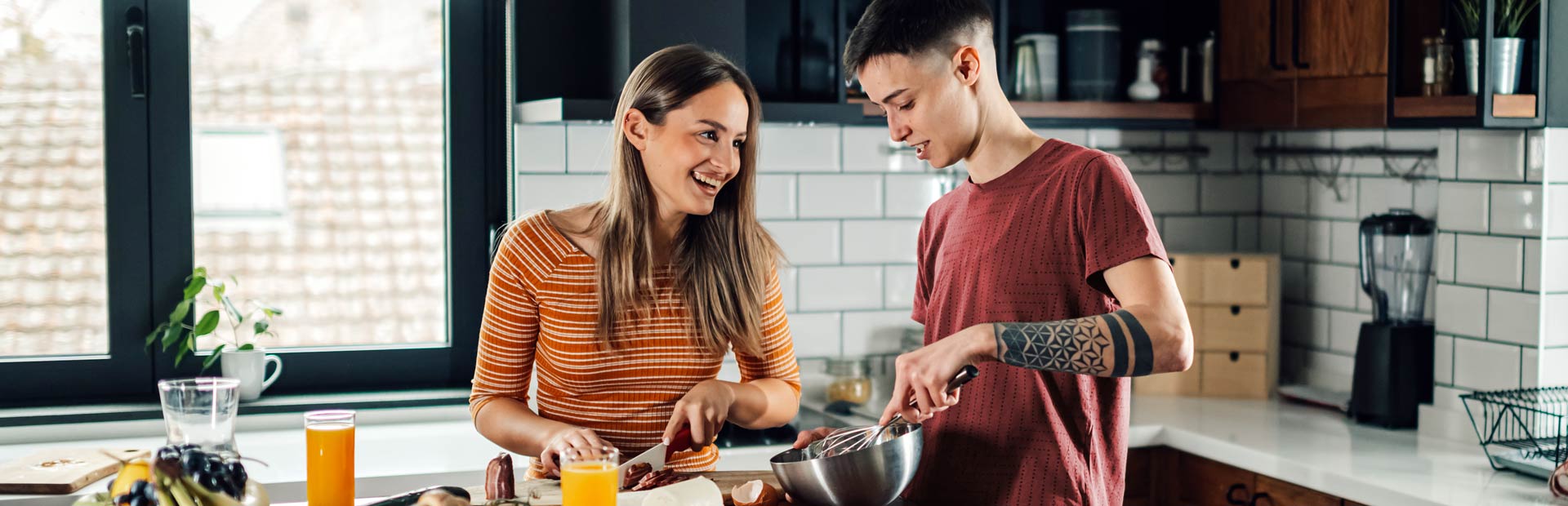 Two people stand comfortably in a kitchen preparing food. One looks down, spreading something with a knife, while the other looks up at them, smiling brightly. The kitchen is sunny, clean, and lived-in.