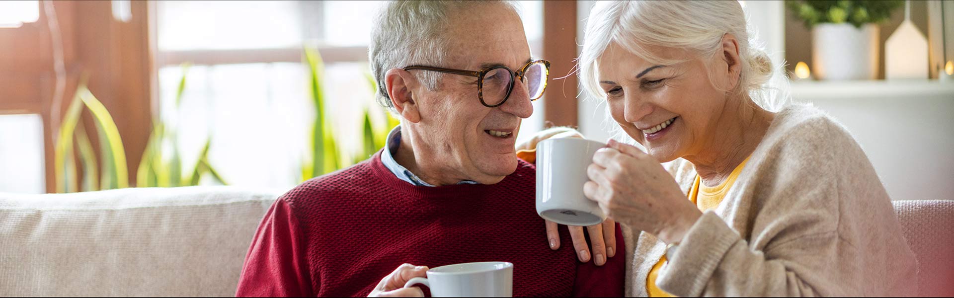 A smiling couple sit side by side on their couch drinking coffee.