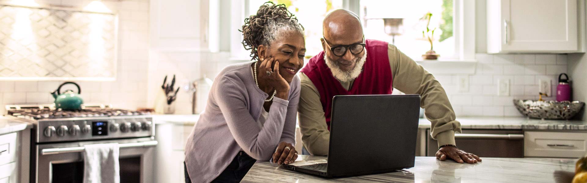 A couple standing in their kitchen looks at investments on their laptop. 