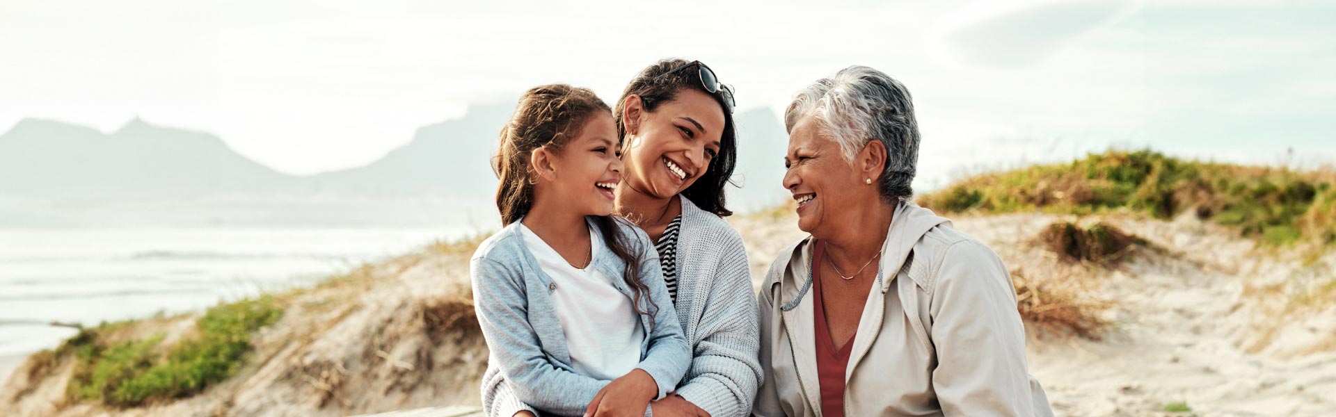  A daughter, a mother, and a grandmother are sitting at the beach, smiling joyfully.