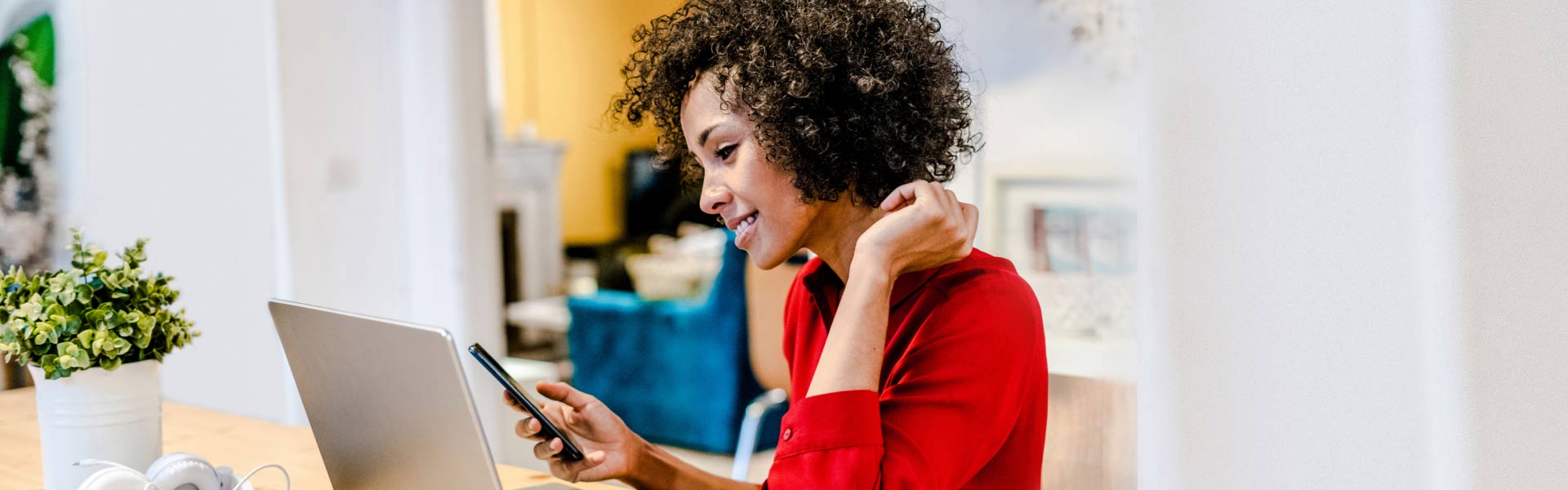 A woman is sitting at a table and smiling as she looks at her phone. She is wearing a red shirt and has curly black hair. There is a laptop in front of her.