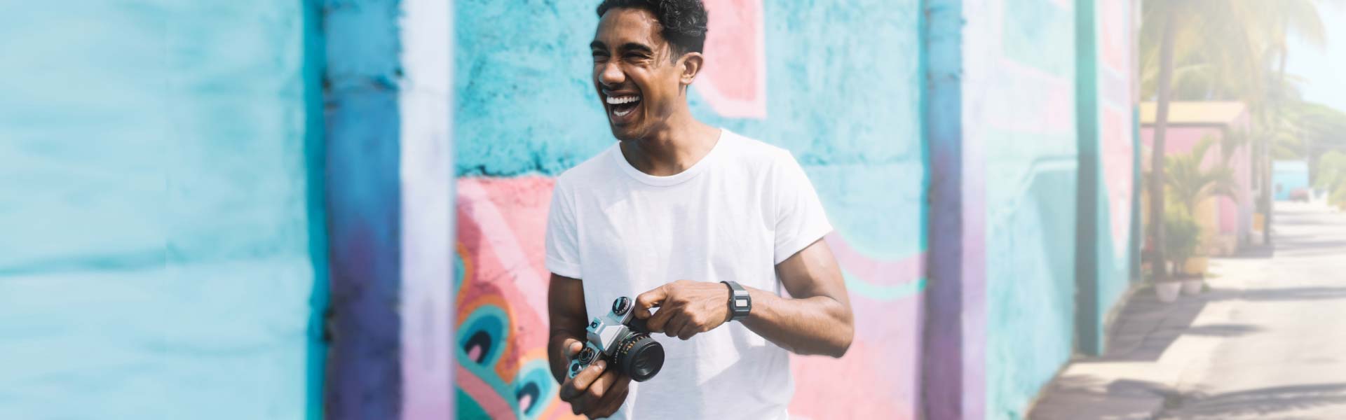 Young man holding a camera and walking down a city street in front of a colorful wall mural.