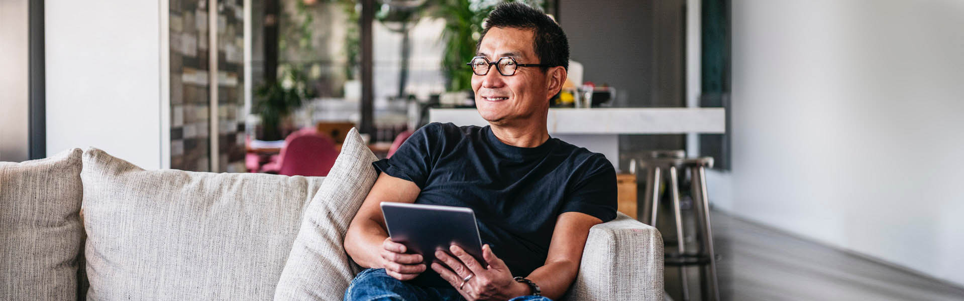 Person with glasses holding an electronic tablet. They are smiling and sitting on a couch in a living space.
