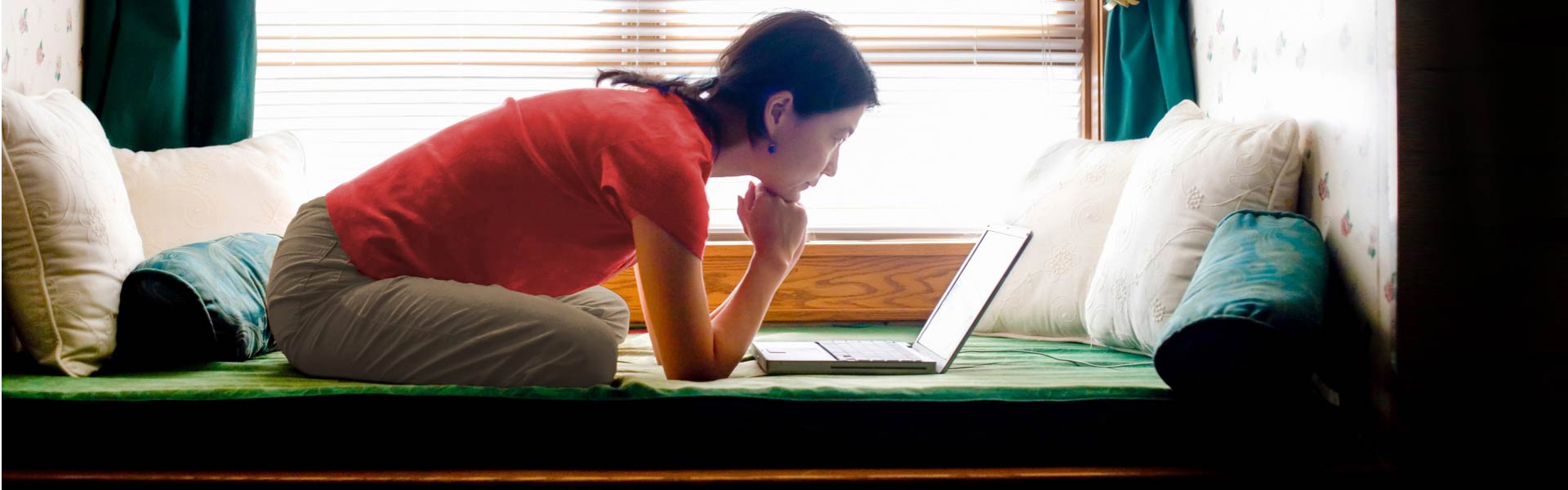 A person sits near a window and leans forward on their elbows while intently looking at their laptop.