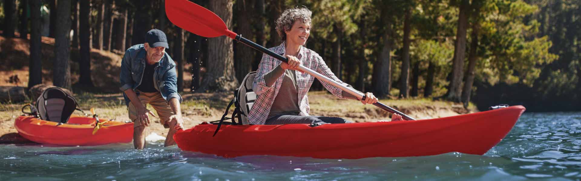 A man and a woman setting off in red kayaks on a river through a forested area.