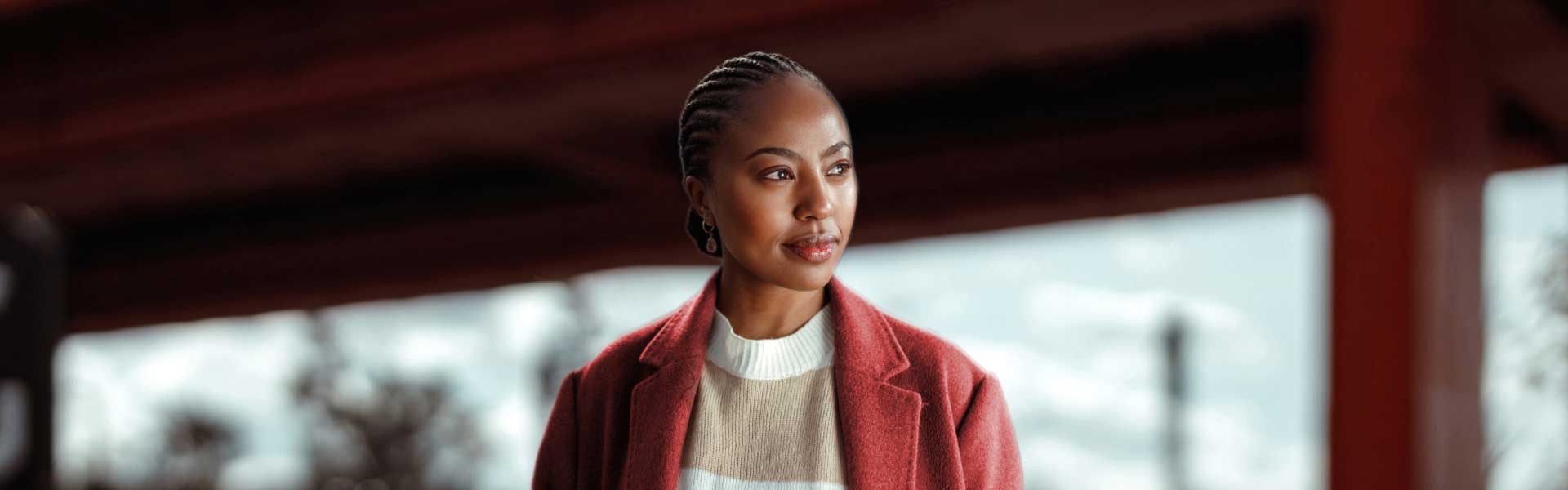 A woman with braided hair, wearing a red wool coat and striped sweater, looks pensively over her left shoulder.