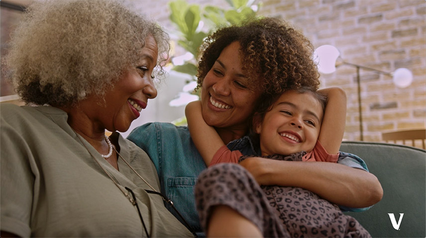 A mother and grandmother are smiling at each other and seated next to each other on a couch, while the mother is tightly holding her smiling daughter on her lap.