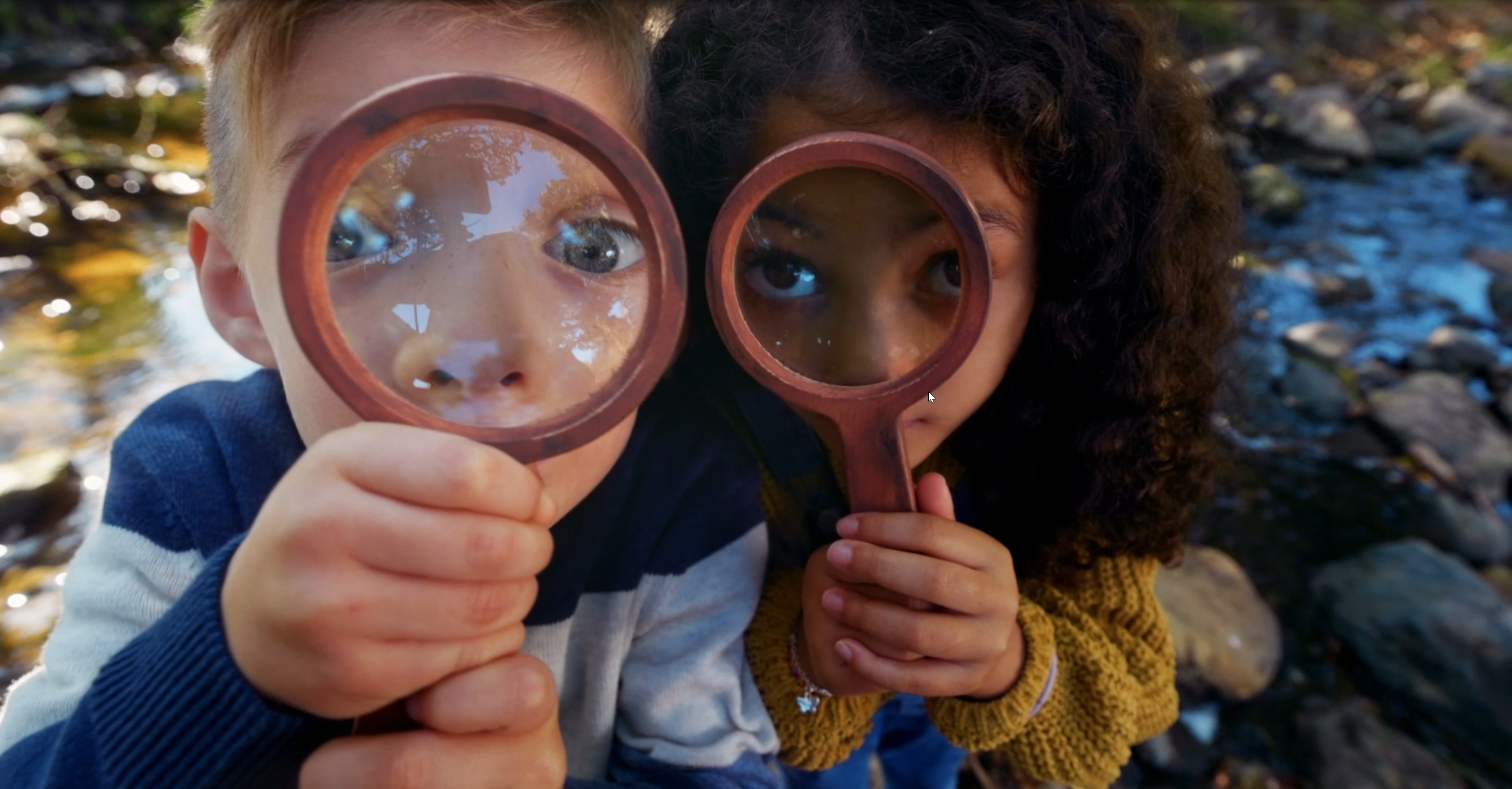 Two kids with magnifying glasses explore a creek bed.