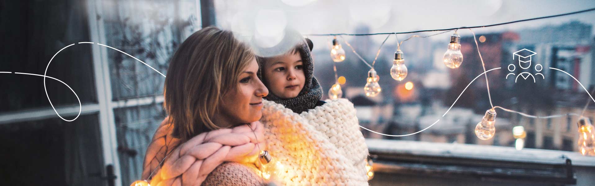Mother holds her son while standing on an apartment balcony overlooking the city. They're wrapped in cozy knits and admiring twinkling string lights.