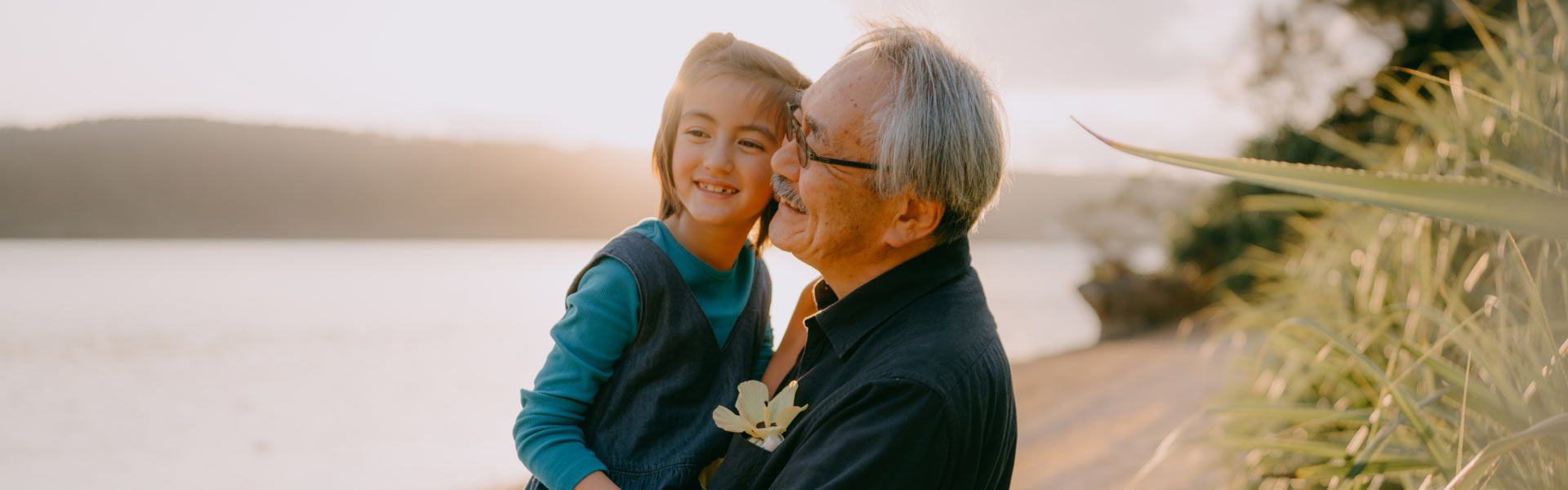 A smiling grandfather holding his happy granddaughter while enjoying the sunset over the water. 