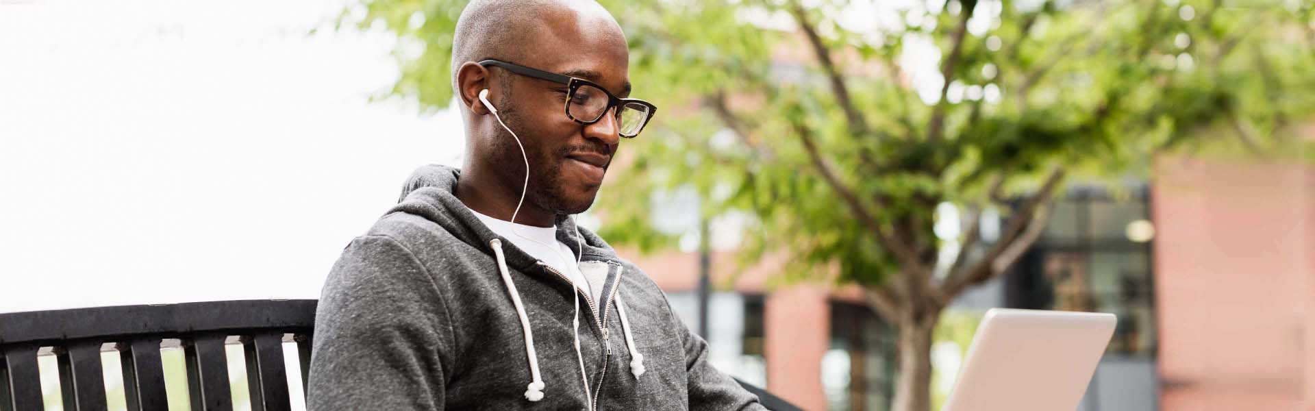 Person using laptop and earphones on city bench.