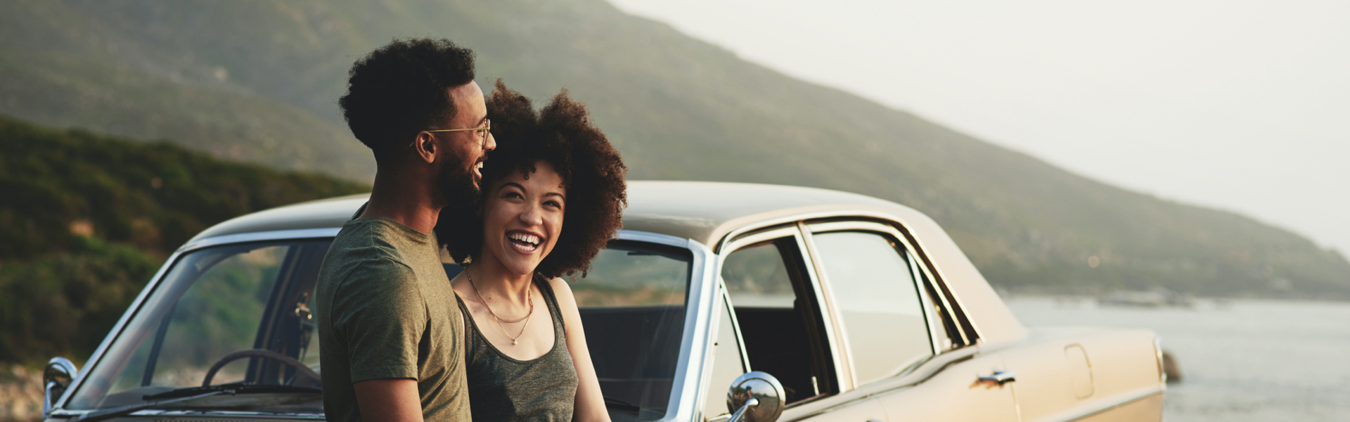 A young couple is smiling while resting against the hood of a car.