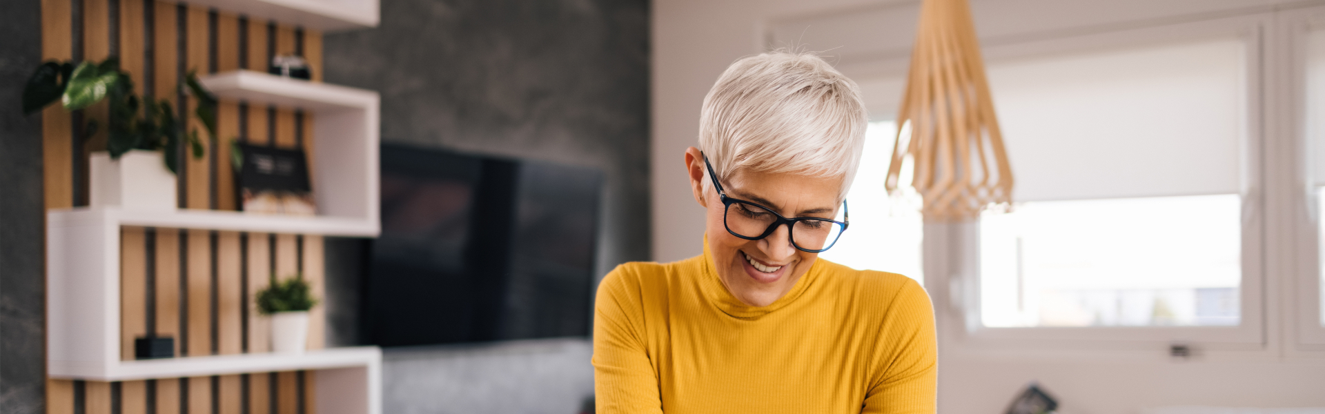 A woman with short blond hair and wearing glasses smiles as she looks down at something out of frame. She is wearing a yellow sweater and is standing in front of a wooden shelf.