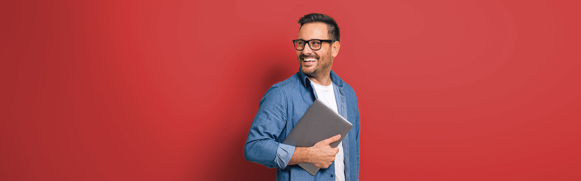 A man wearing a blue shirt and glasses smiles and holds a laptop in front of a red background.