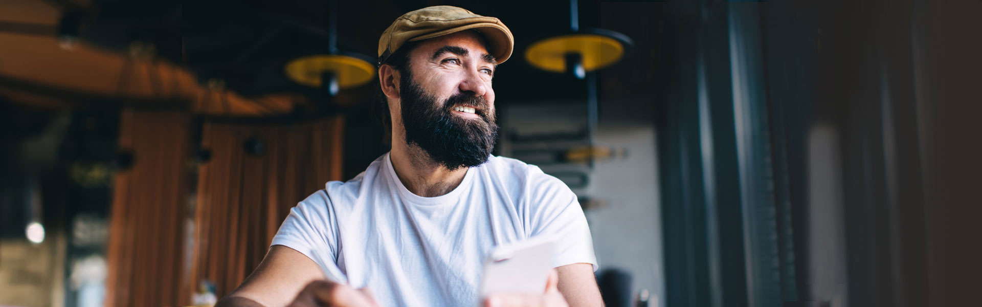  A bearded man wearing a white shirt and a pageboy cap smiles as he holds his mobile phone.