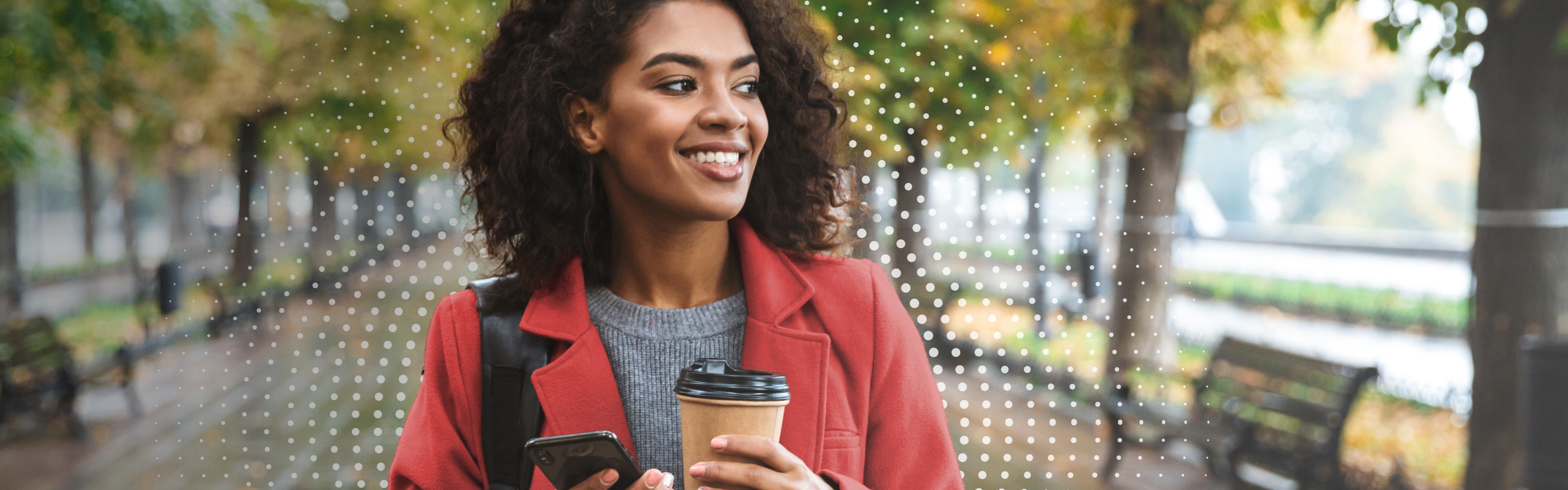 Smiling woman walks along a tree-lined street with phone and coffee in hand.