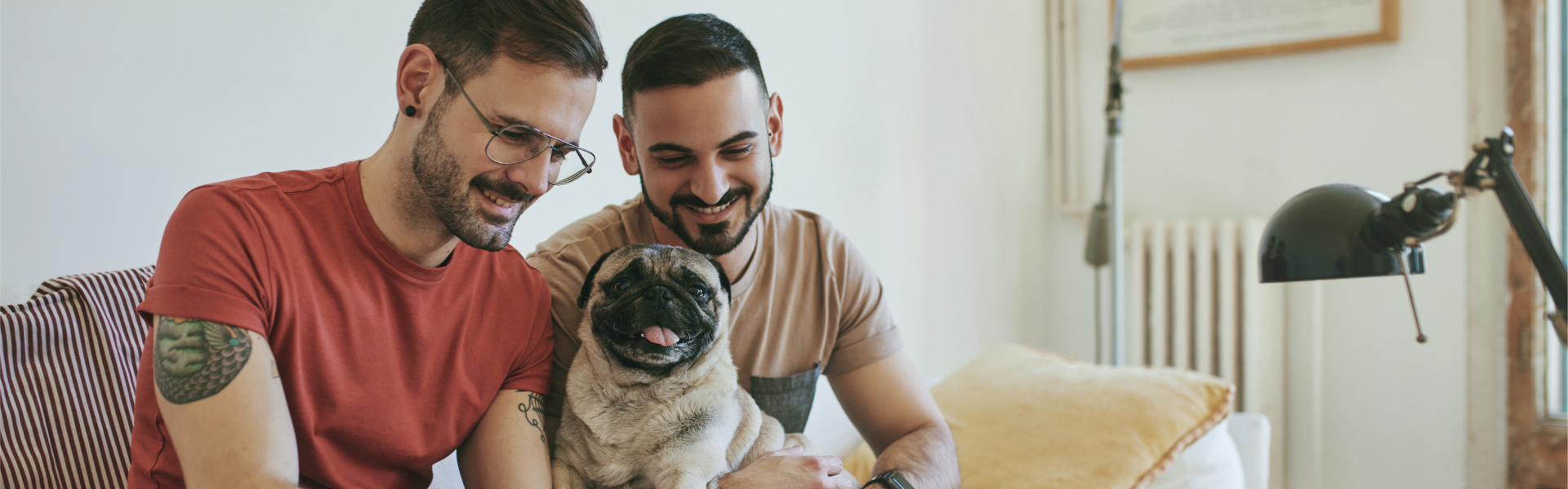 Pair of younger men, with tattoos, sit on sofa with their dog. 