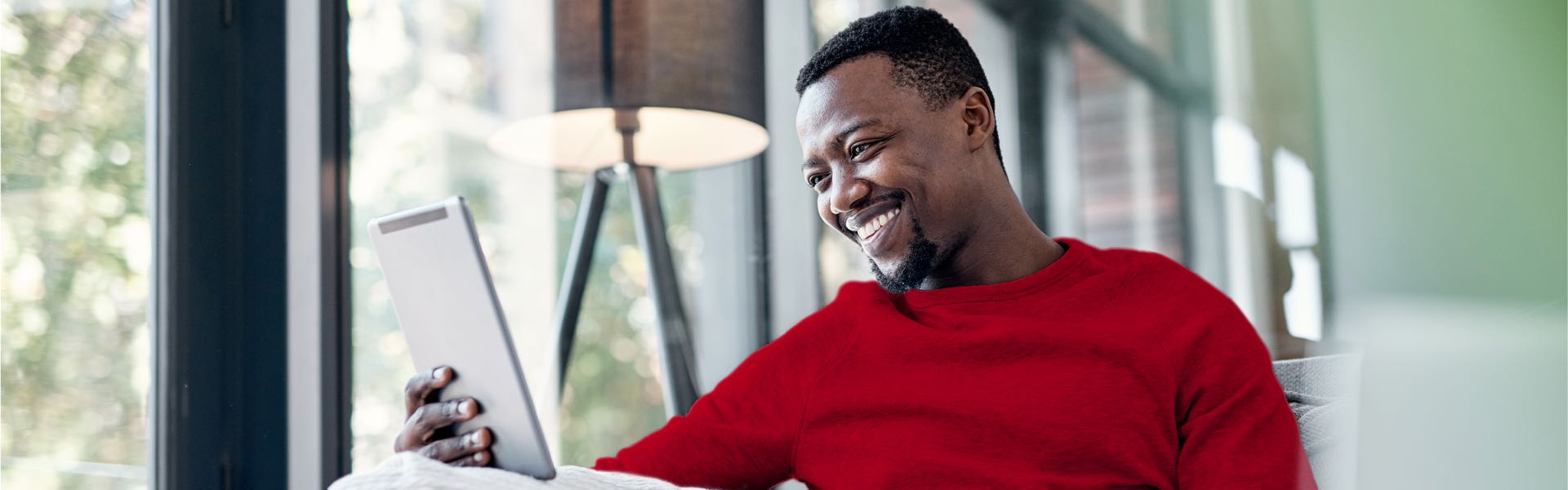 A young man sitting inside, holding an iPad and smiling at it.