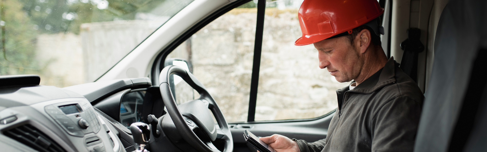 A man wearing hard hat is checking his phone in his work van.