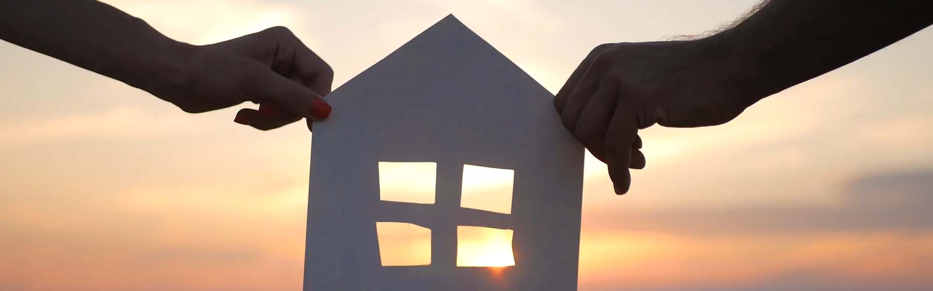 Two hands holding a cut out of a paper house against a sunset.