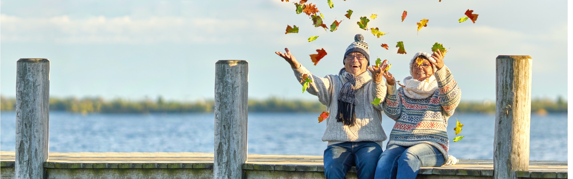 An elderly couple sitting on a dock is enjoying the fall weather and joyously throwing up leaves while bundled in cozy sweaters.	