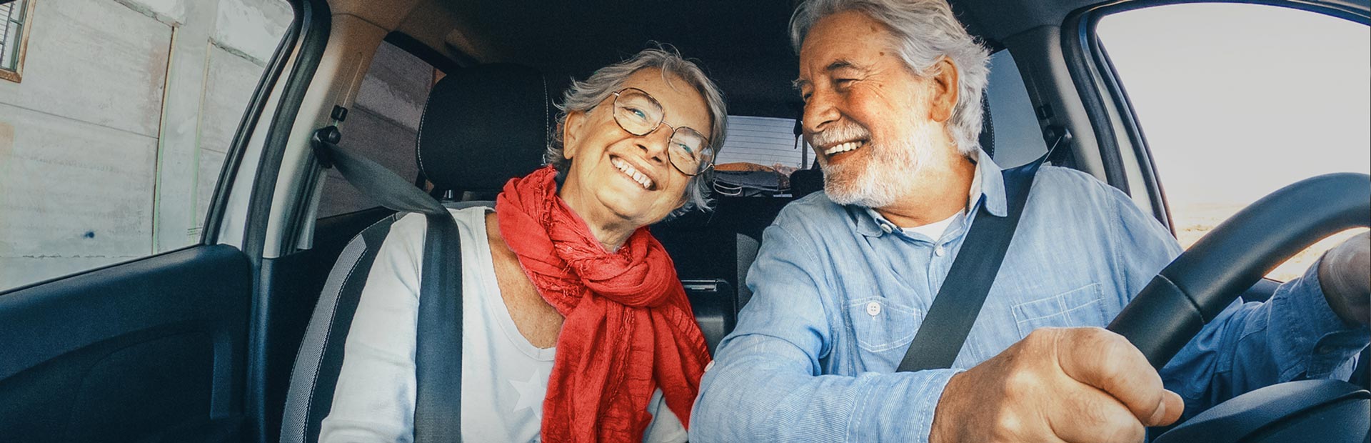 =An older couple in the front seat of a car are smiling with anticipation, like they’re about to embark on an adventure. 