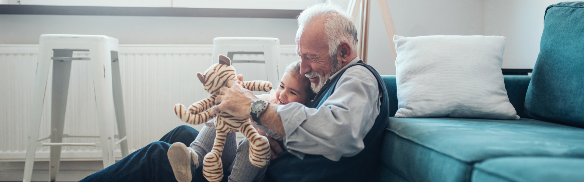  While seated on the floor in front of a couch, a smiling grandfather plays with a stuffed animal with his granddaughter seated in his lap.