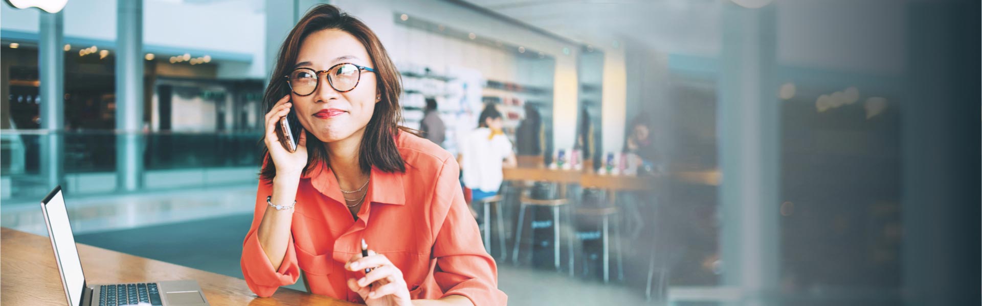 A young woman with round glasses sits with her laptop in a restaurant while talking on the phone.