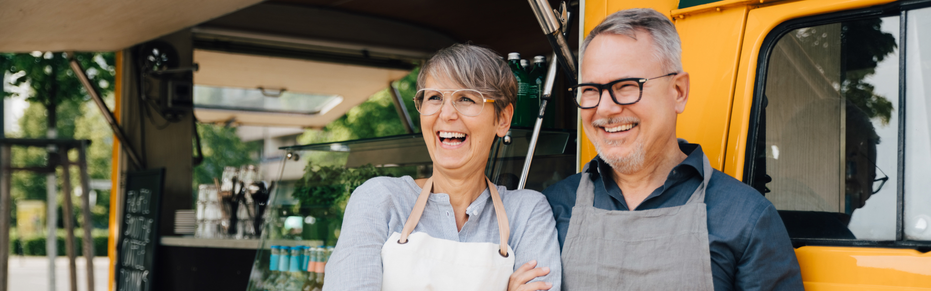 An older man and woman are smiling and standing in front of a food truck.