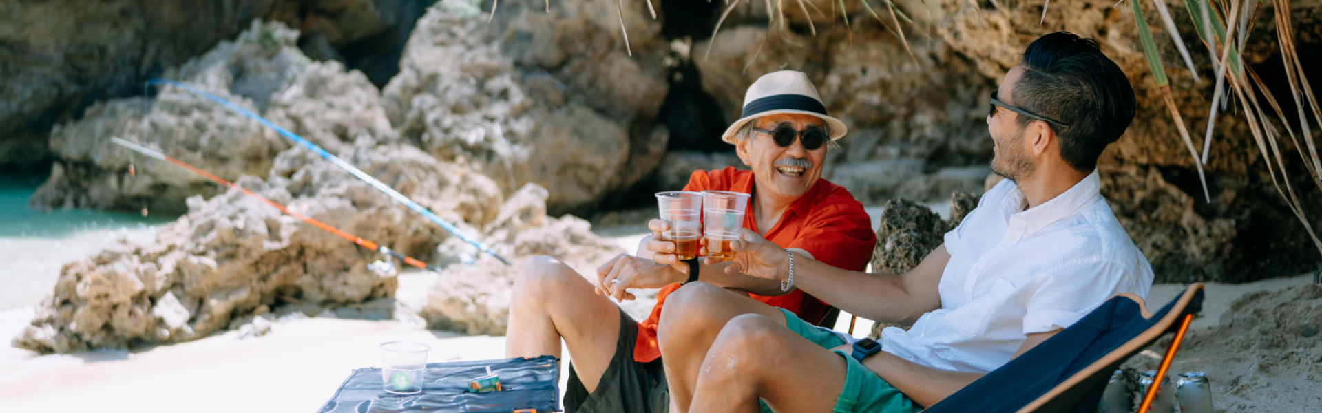 Two men are sitting on chairs on a beach with drinks in their hands.