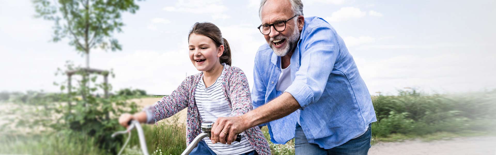 A father and daughter are outside. The daughter is riding a bike and her father is helping to guide her by holding the handlebars. 