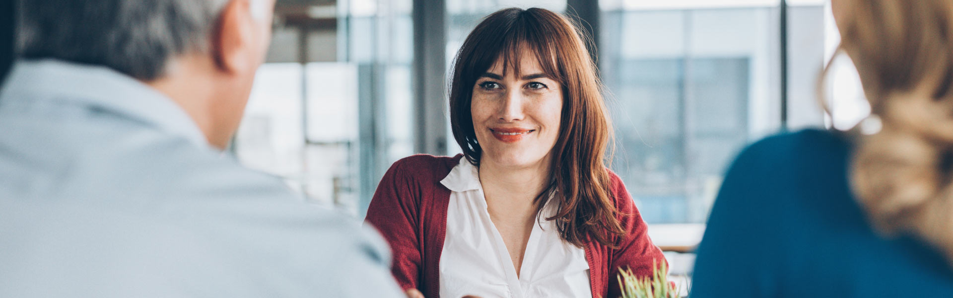 A woman wearing a white shirt and a red sweater is smiling, sitting in an office across from two other people.