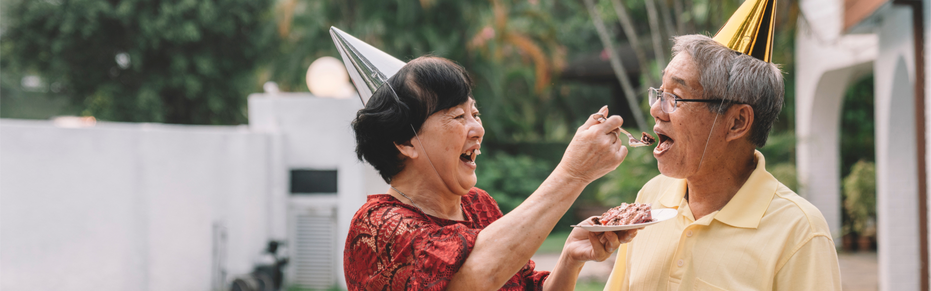 Mature, retirement age couple celebrating birthday with party hats on. The lady is laughing and feeding her husband cake.	
