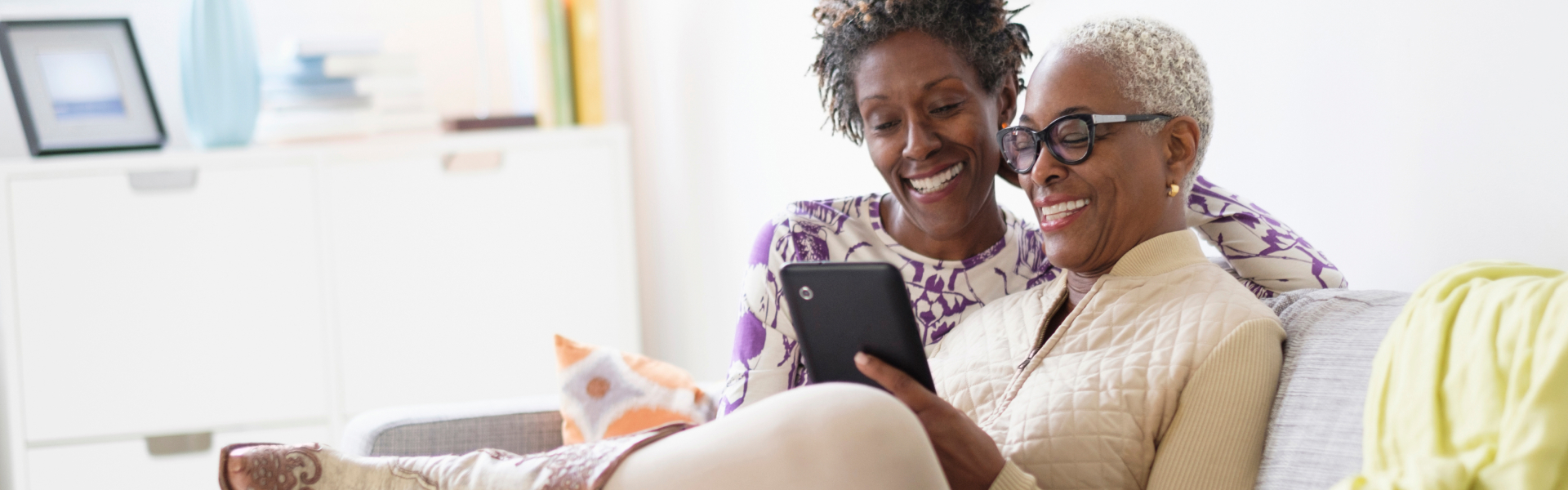 Two women sitting on a couch looking at an iPad and smiling.