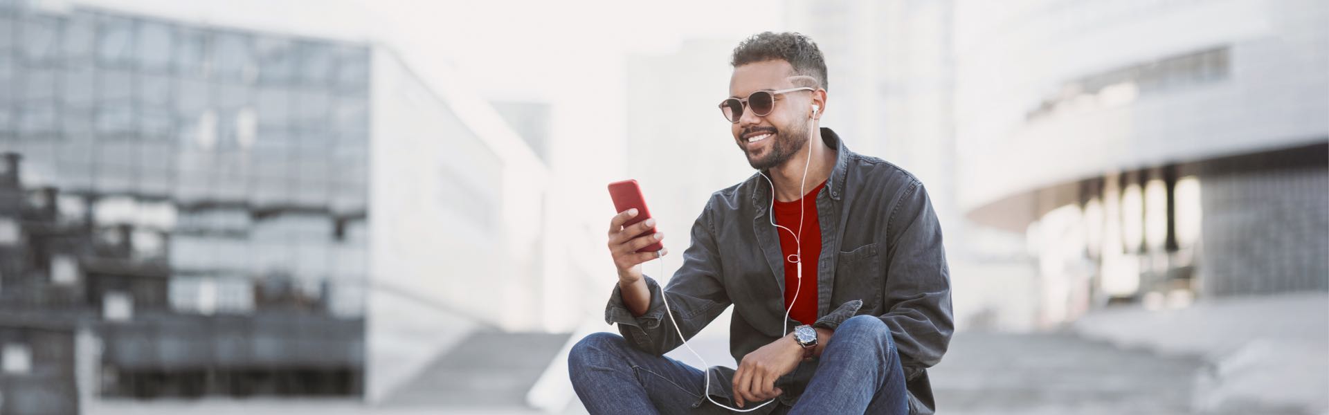 Smiling man in sunglasses and dark jacket using red smartphone while sitting outdoors in a urban setting.