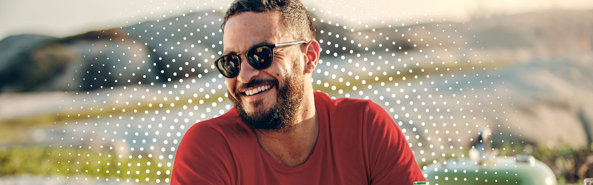 A young man sitting outside with sunglasses on, smiling and laughing.