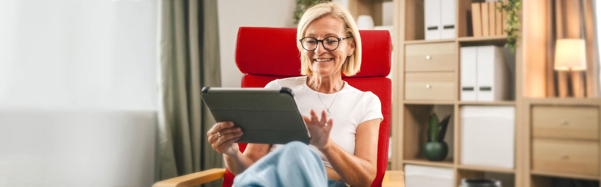 Woman sitting in a red chair smiles as she reads something on a tablet.