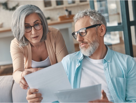 An older man is sharing documents with an older woman who is learning over his shoulder to read them.