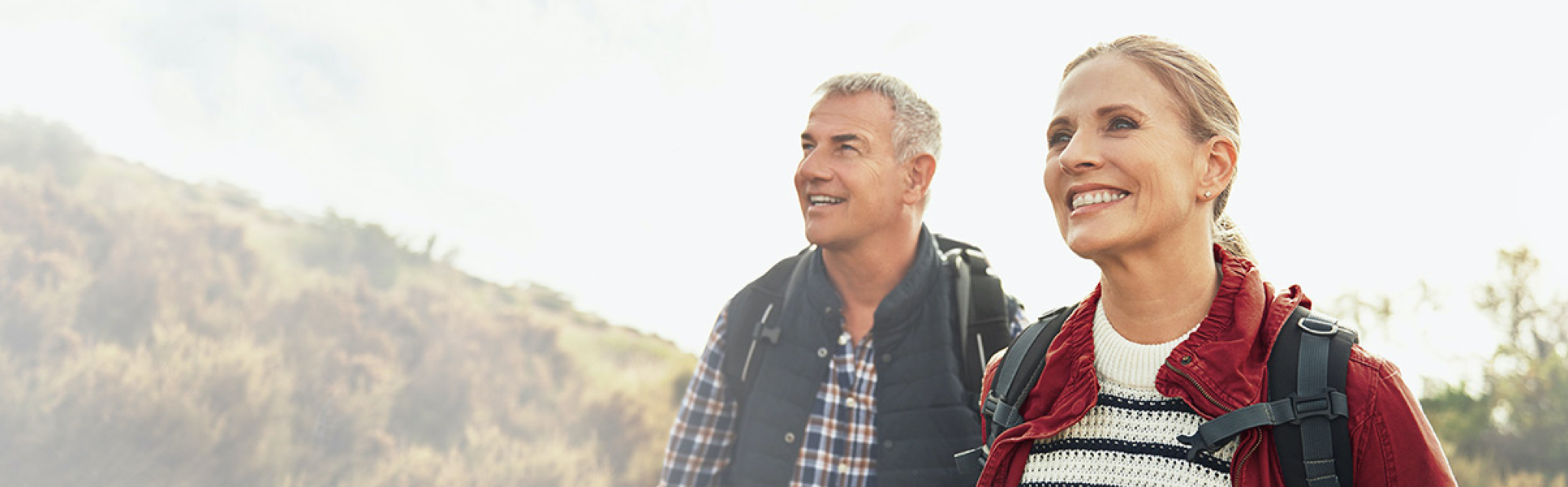 An older couple (man and woman) hiking outside with backpacks on and smiling.