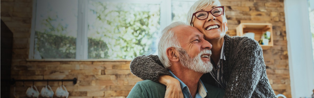 A woman leans over to embrace her husband in their kitchen. 