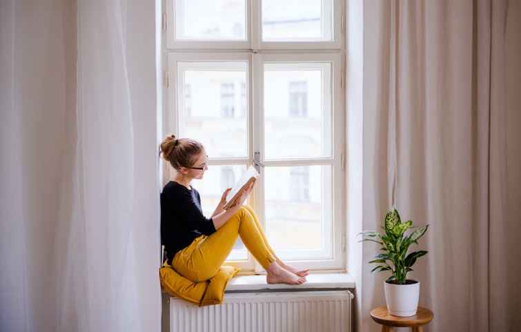 Woman sitting on a yellow pillow in a window seat, relaxing and enjoying a book.