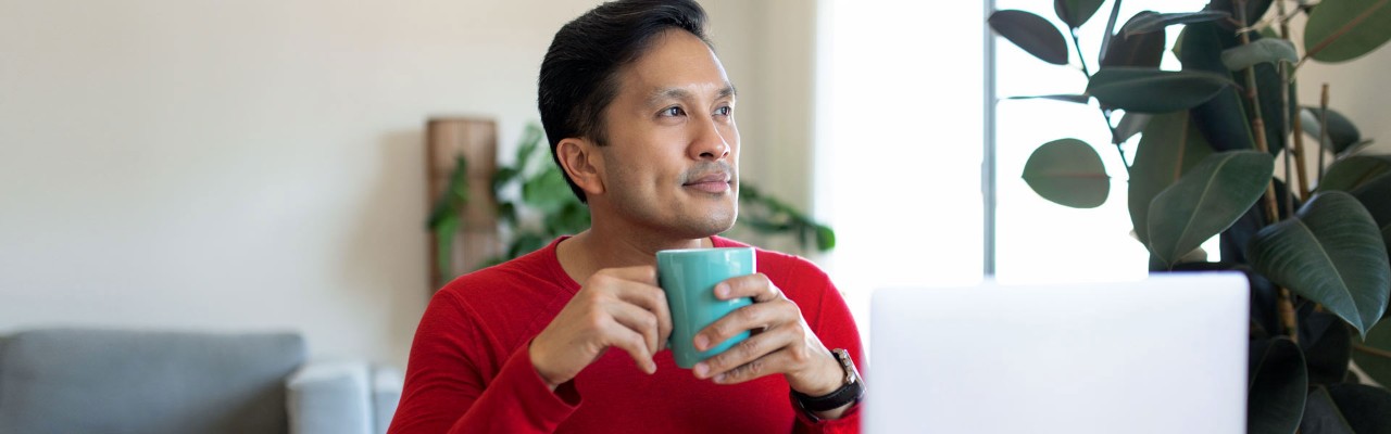  A man holding a mug with both hands is smiling and looking out into the distance on his left.