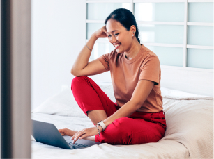 A young woman sitting on a bed is resting her arm on her knee and smiling down are her open laptop.