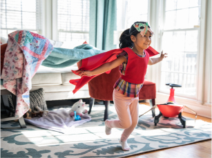 A young girl wearing a cape on her back and a butterfly mask on her head is smiling while running forward with her arms stretched out.