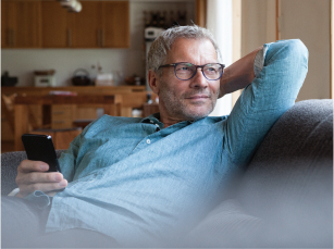  A middle-aged man holding a smartphone is wearing glasses while sitting on a couch with his arm tucked behind his head while he looks out to his left.