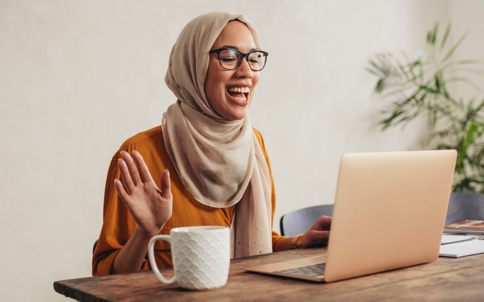  A young woman wearing a hijab and glasses is openly smiling at a laptop screen while open-palm gesturing with one of her hands.