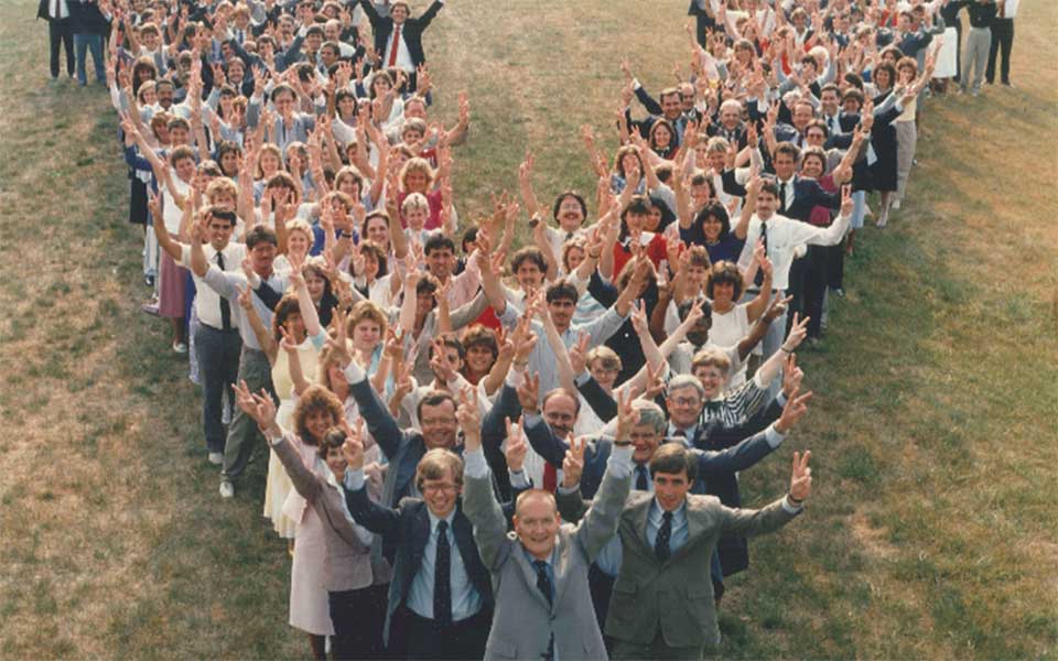 Photo of a large group of businesspeople organized into the letter V, smiling and holding up two fingers.