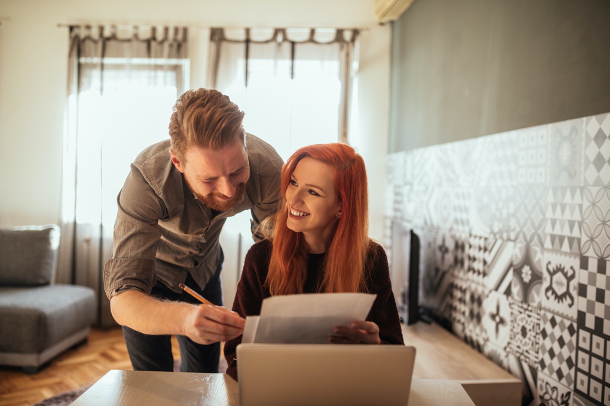 Seated woman with a man standing over her shoulder, looking at account transfer paperwork in their living room. 